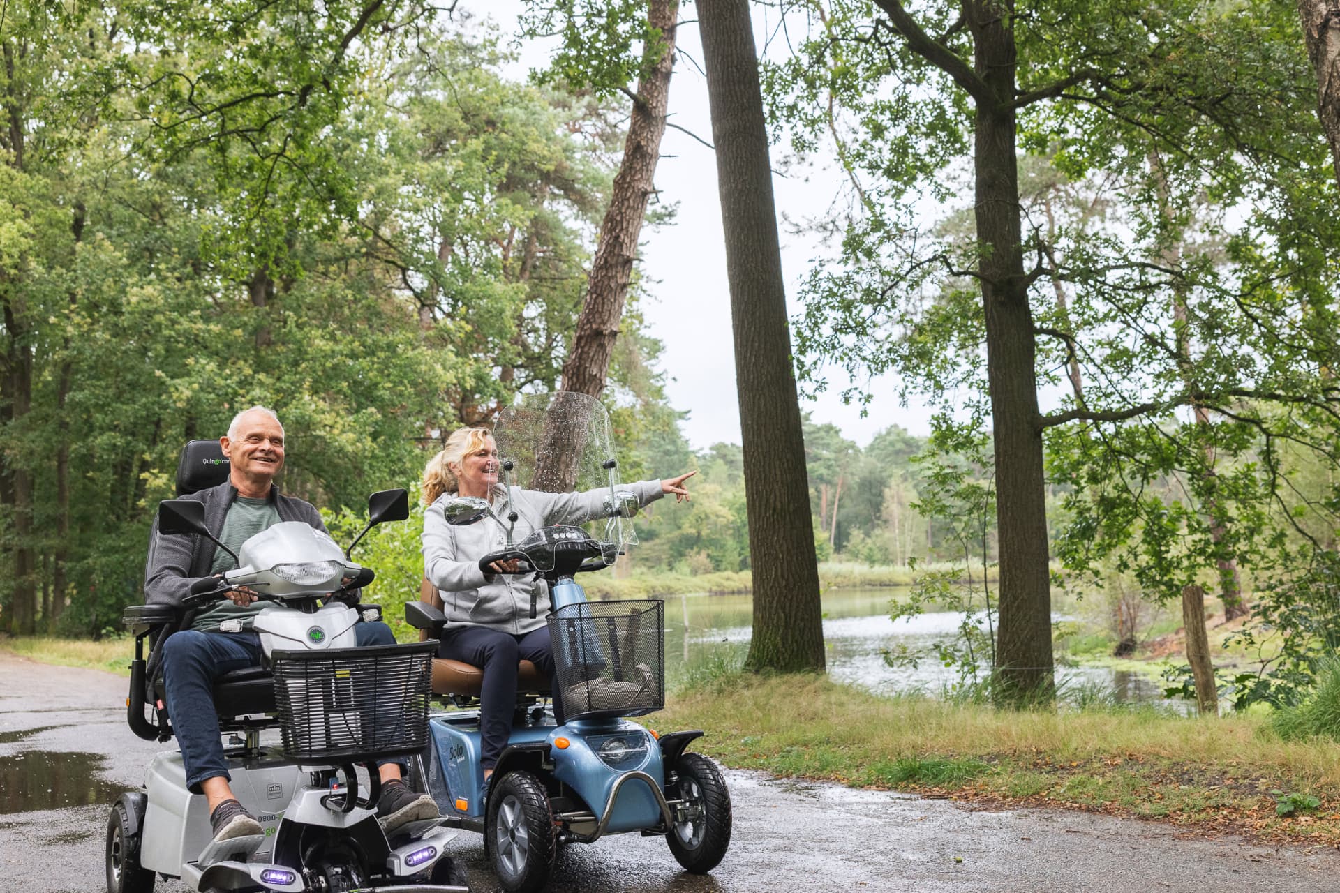 Twee mensen rijden samen op scootmobielen langs een meer.
