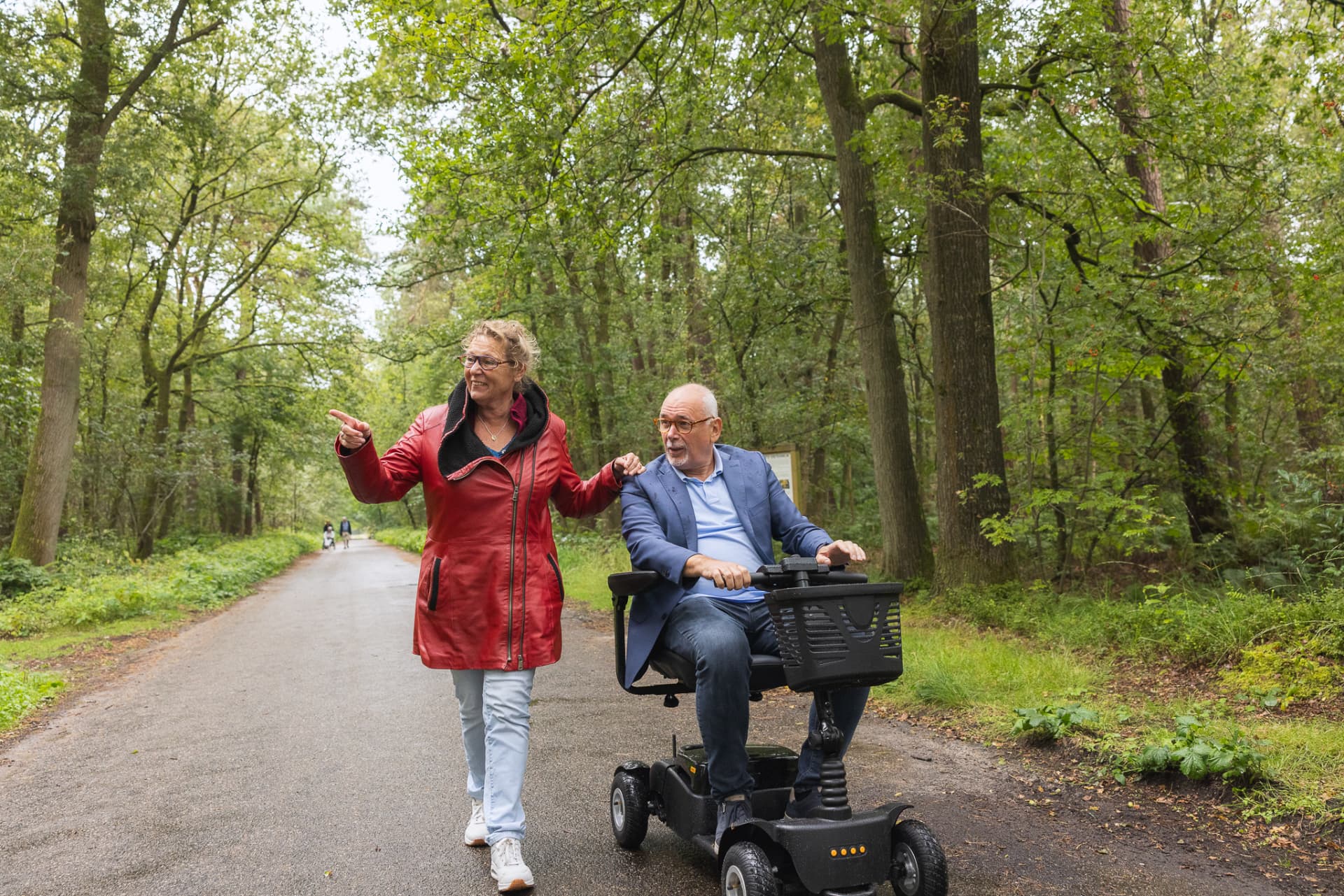 Een vrouw loopt naast een man op een scootmobiel over een bosweg.