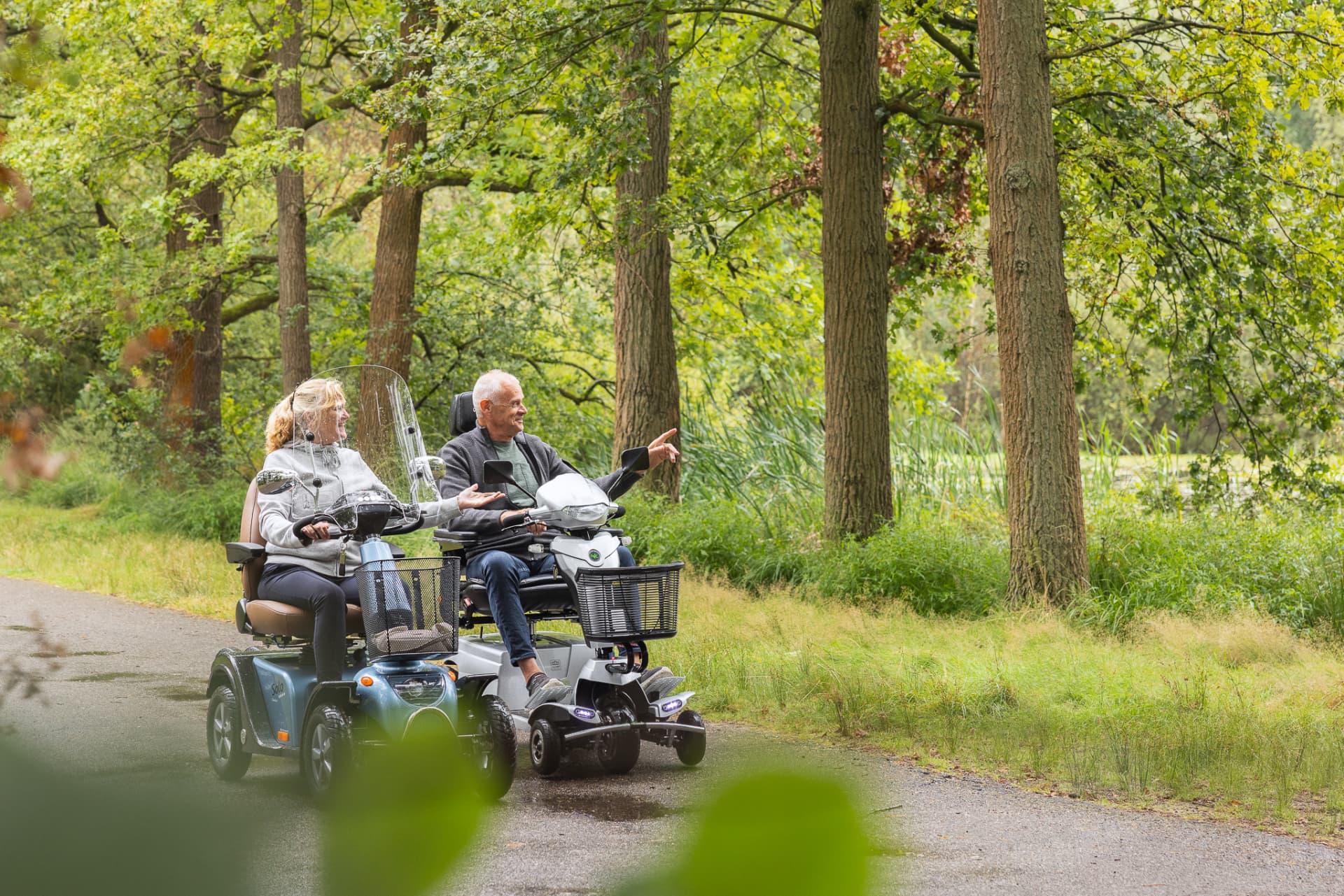 Twee mensen rijden naast elkaar op scootmobielen over een bospad.